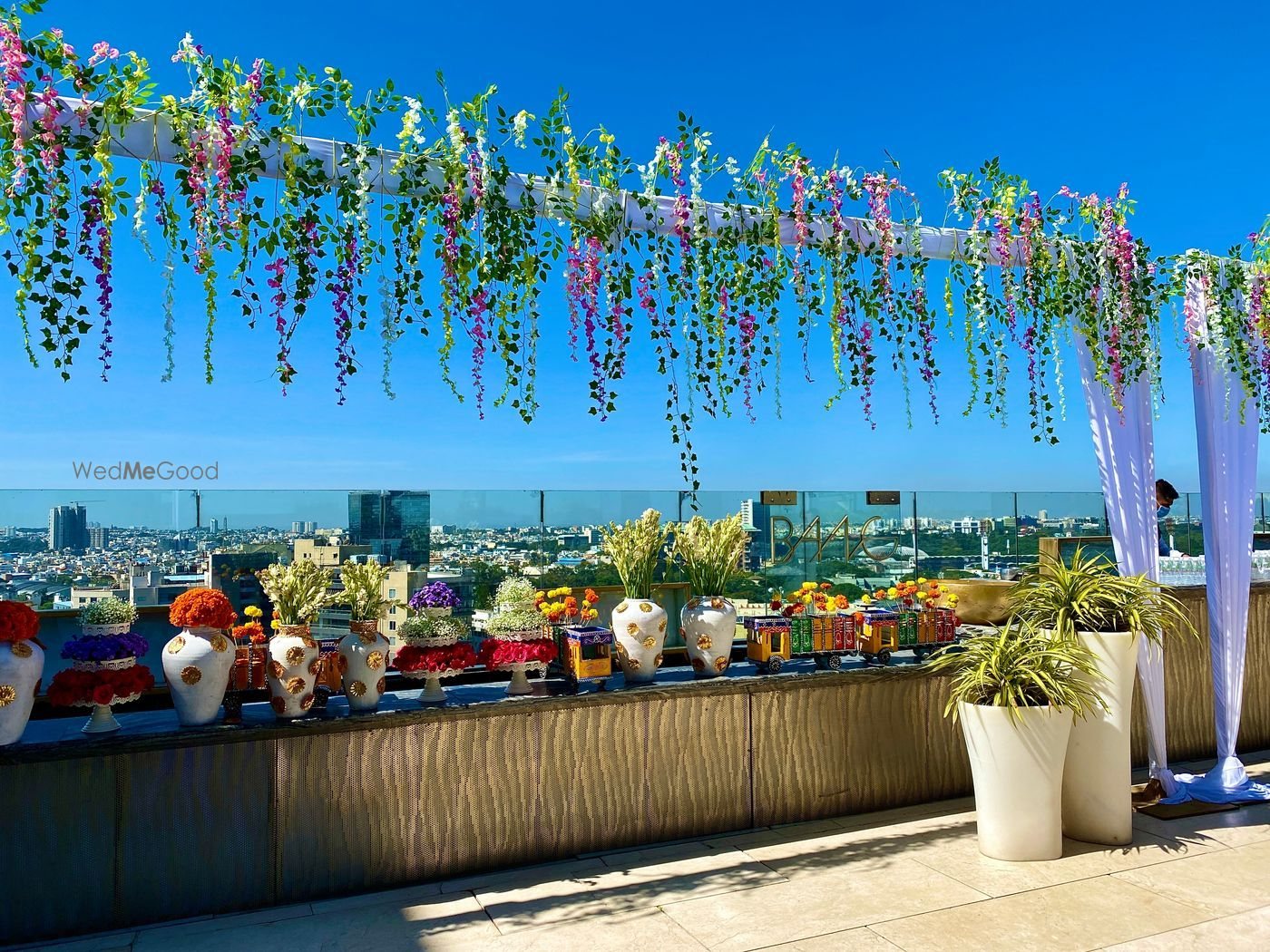 BANG, the rooftop terrace at The Ritz-Carlton Bangalore, dressed for a daytime function with a hanging canopy of trailing greenery and orchids over a long console of marigold and tuberose urns, with the city skyline behind the glass balustrade