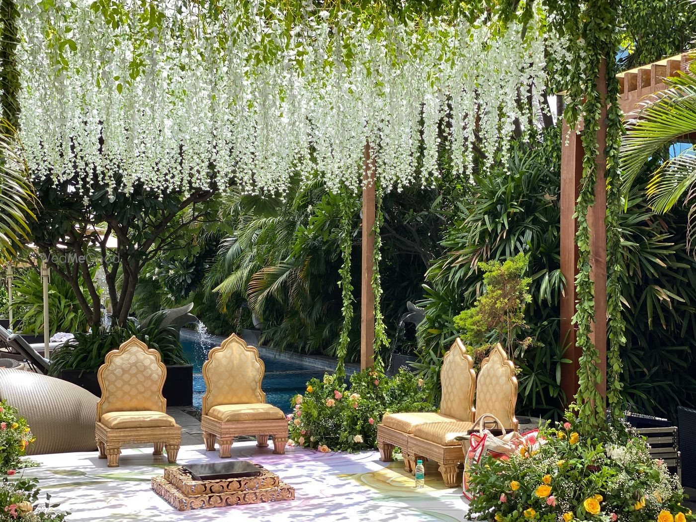 A ceremony platform set beside the outdoor pool at The Ritz-Carlton Bangalore under a dense hanging canopy of white blossom, with gilded low seating, a fire vessel and tropical planting behind