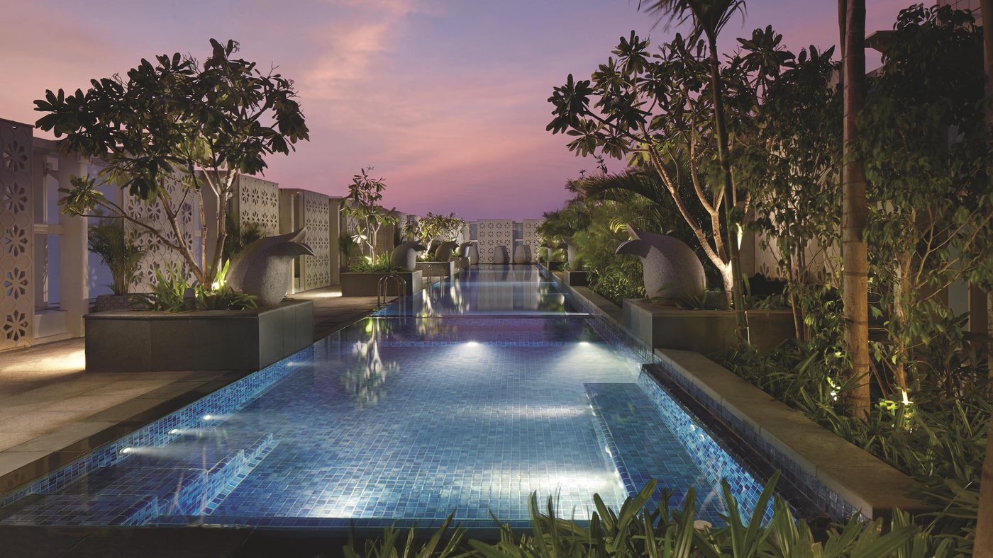 The outdoor pool at The Ritz-Carlton Bangalore at dusk — a mosaic-tiled lap pool lit from within, flanked by perforated jaali screen walls, stone bird sculptures and frangipani trees under a pink sky