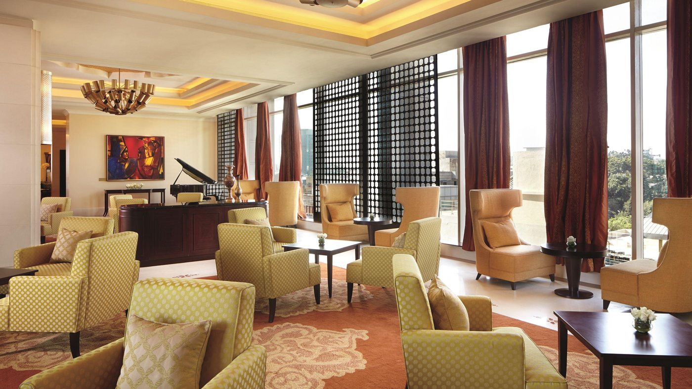 The lobby lounge at The Ritz-Carlton Bangalore — gold and citron armchairs on a patterned carpet, a grand piano, and full-height windows behind dark perforated jaali screens and red drapes