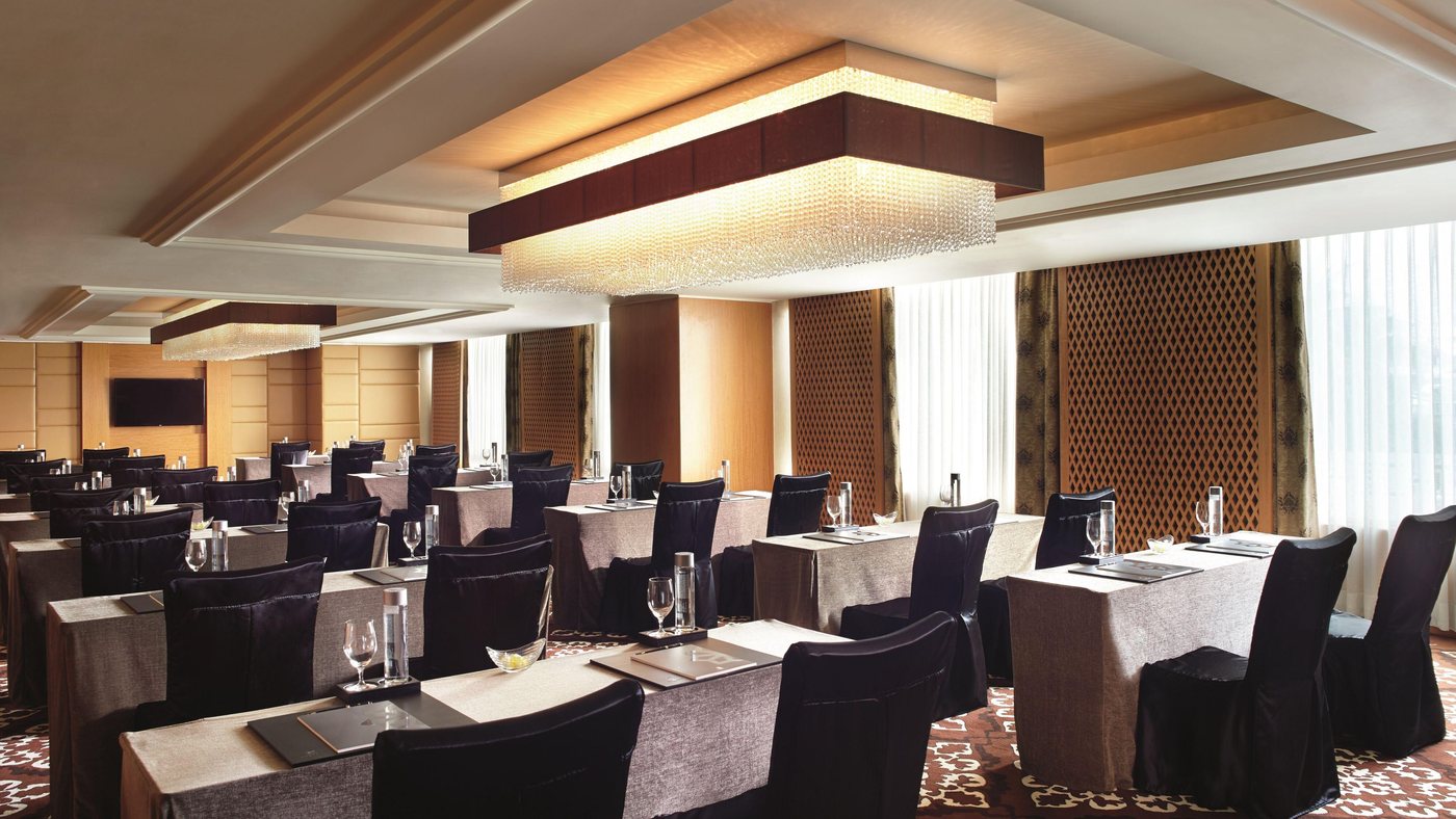A function room at The Ritz-Carlton Bangalore laid out classroom-style under two rectangular crystal-strand ceiling fixtures, with wood-panelled walls and diamond-patterned screens