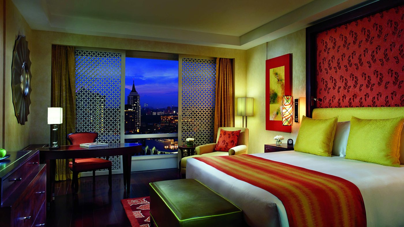 A guest room at The Ritz-Carlton Bangalore at dusk — patterned headboard panel, jaali-screened window and a desk, with the lit city skyline beyond