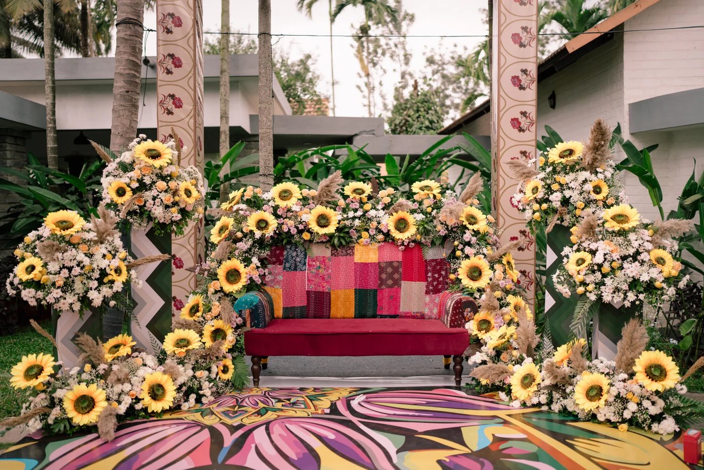 A mehendi seat dressed on the lawn beside Samaya's white guest cottages — a crimson settee against a patchwork kantha backdrop, sunflower and pampas towers either side, a painted floral floor and pastel drapes stretched overhead between block-printed pillars
