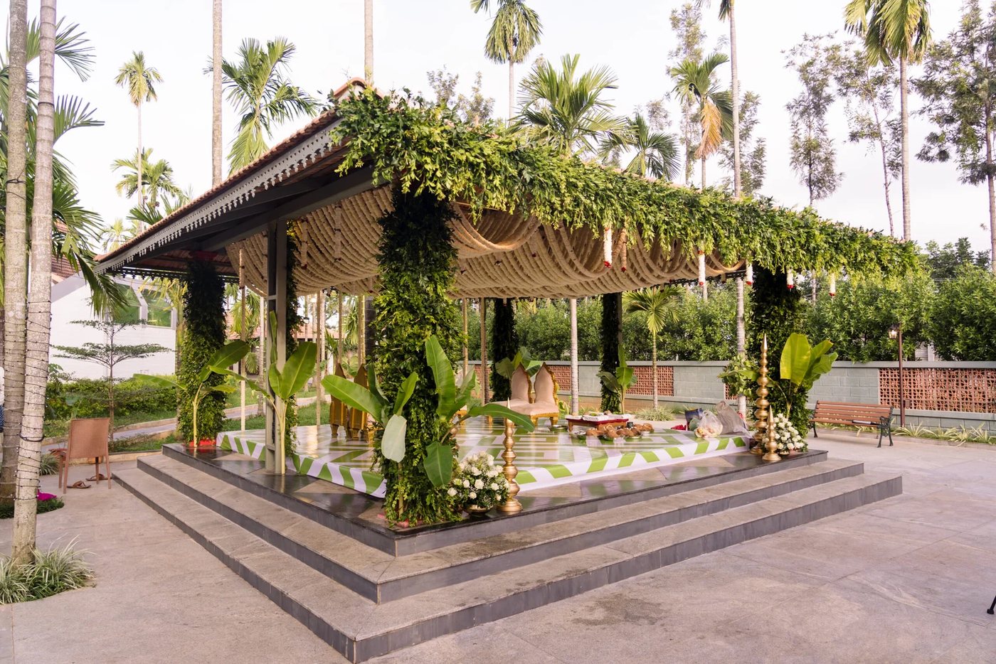 An open mandap built on the raised granite plinth of the Kalasha Outdoors pavilion in late-afternoon light — rope swags looped under the tile roof, columns wrapped in foliage, banana stems at the corners and a green-and-white geometric floor, with areca palms behind