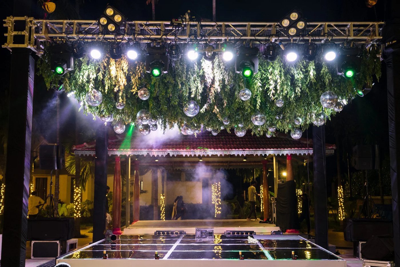 Sangeet night at Samaya — a ground-supported lighting truss carrying moving heads and a foliage-and-mirrorball ceiling rigged in the forecourt in front of the Kalasha pavilion, haze in the beams and a mirrored dance floor laid over the forecourt stone