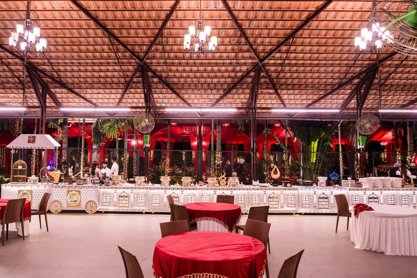 Thambula Dining at night — clay tiles on exposed steel trusses with chandeliers and ceiling fans between the frames, mesh side walls open to the lit plantation, a long white-and-gold buffet counter down one side and round tables laid in red