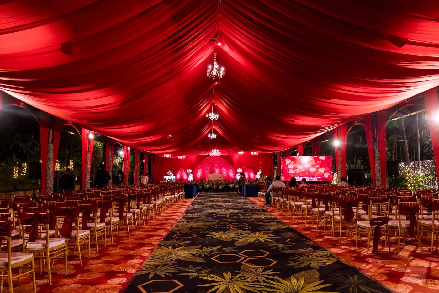 The Kalasha pavilion under a full red fabric ceiling drape for a reception — chandeliers along the ridge, gold chiavari chairs in rows either side of a patterned centre aisle, an LED wall to the right and a rose-wall stage at the far end