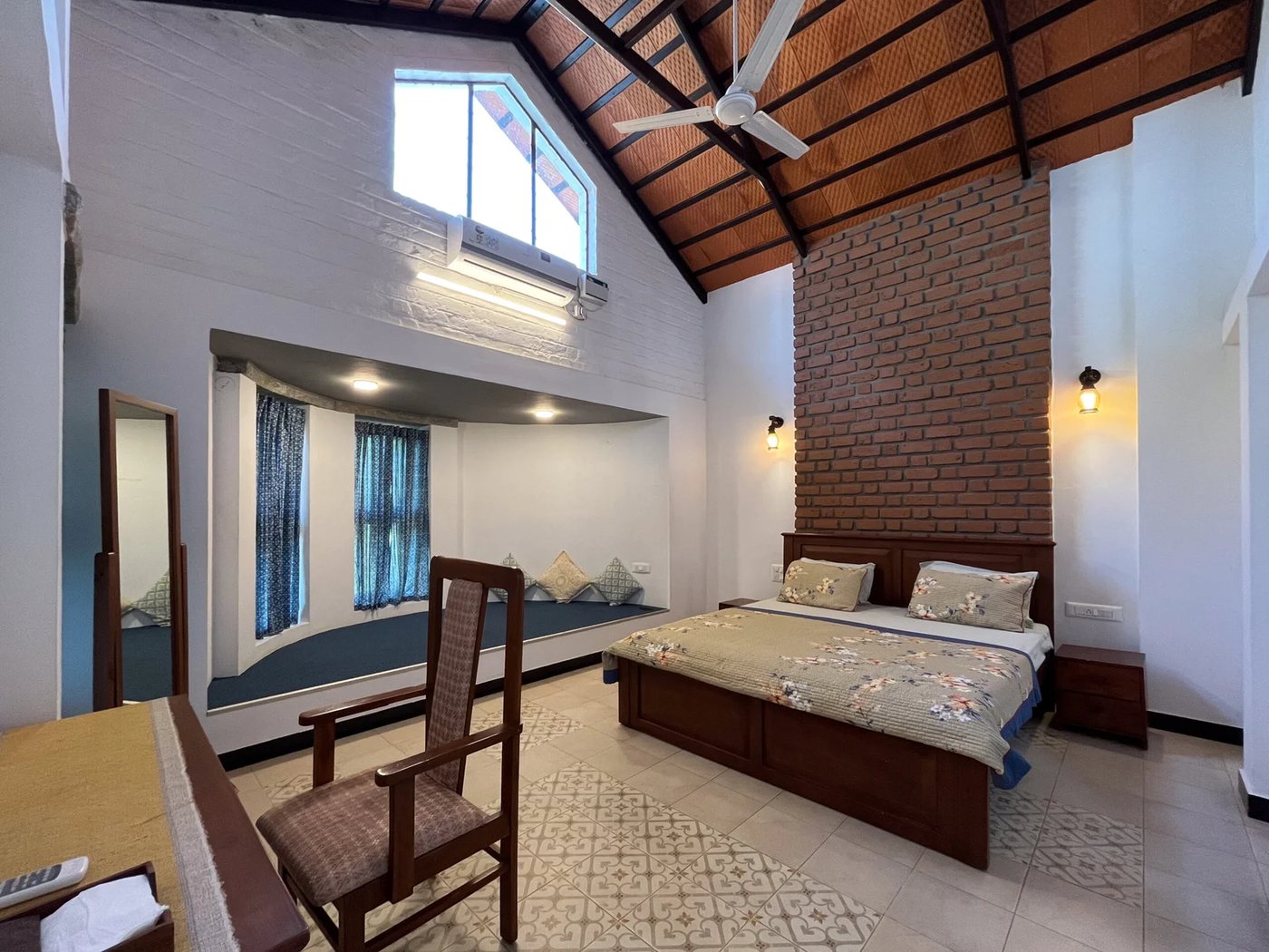 A bedroom in the Hombale House — double-height vaulted ceiling of clay tiles on dark steel rafters, an exposed brick headboard wall, a curtained window seat in the alcove, patterned cement-tile floor, ceiling fan and a wall AC above the clerestory window