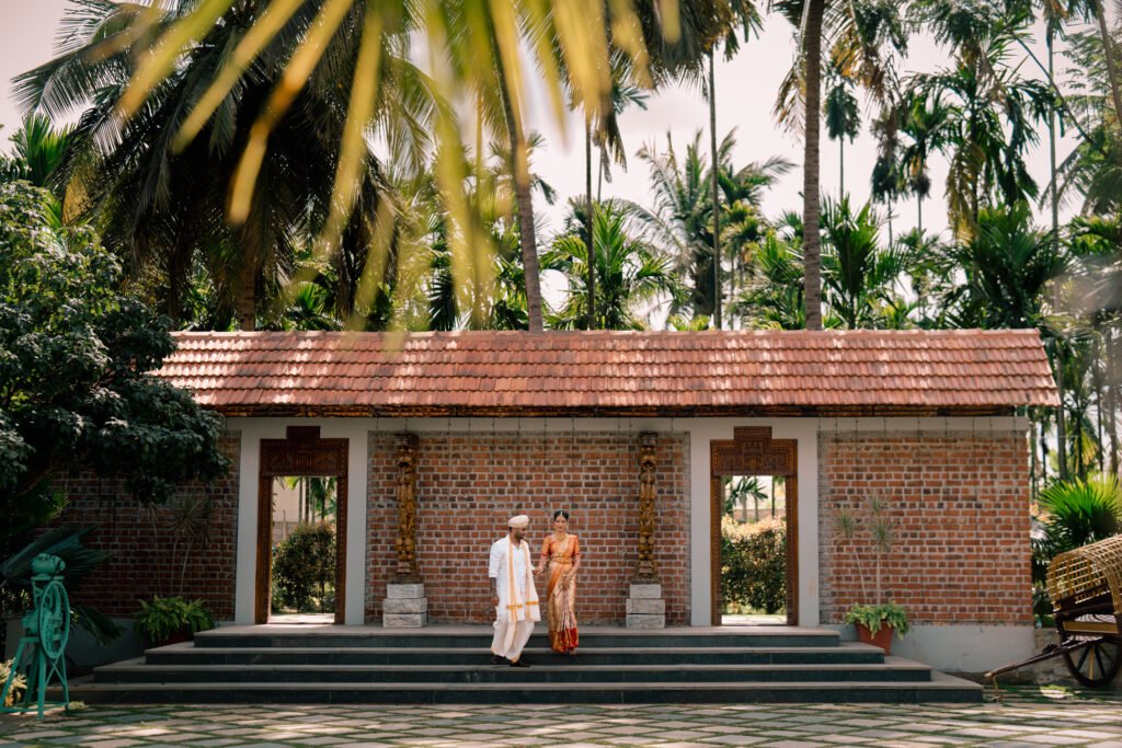 A couple in South Indian wedding dress on the granite steps of Samaya's brick-and-clay-tile guest pavilion, carved teak doorframes either side of the brick wall, tall areca and coconut palms overhead in flat late-morning light