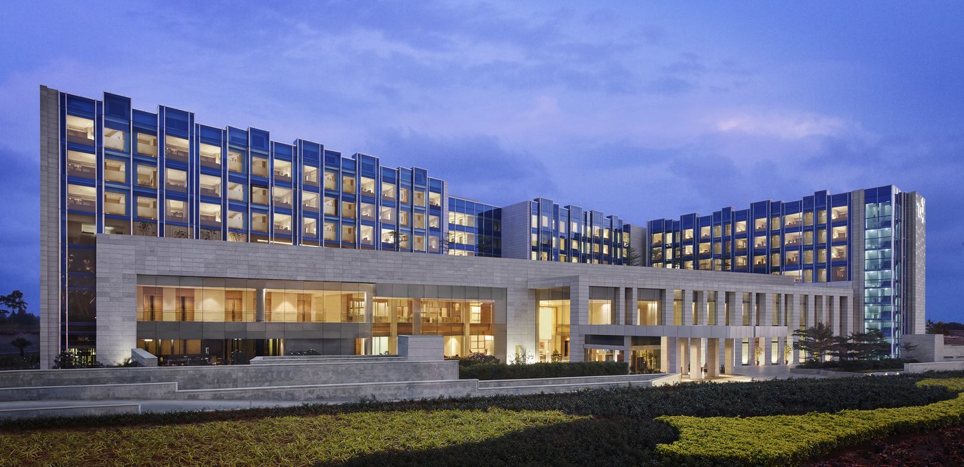 Taj Bangalore at dusk from the garden side — a long low-rise stone and blue-glass hotel with a projecting glazed lobby wing and lit guest-room floors stepping up behind, above clipped hedges and a lawn in the foreground