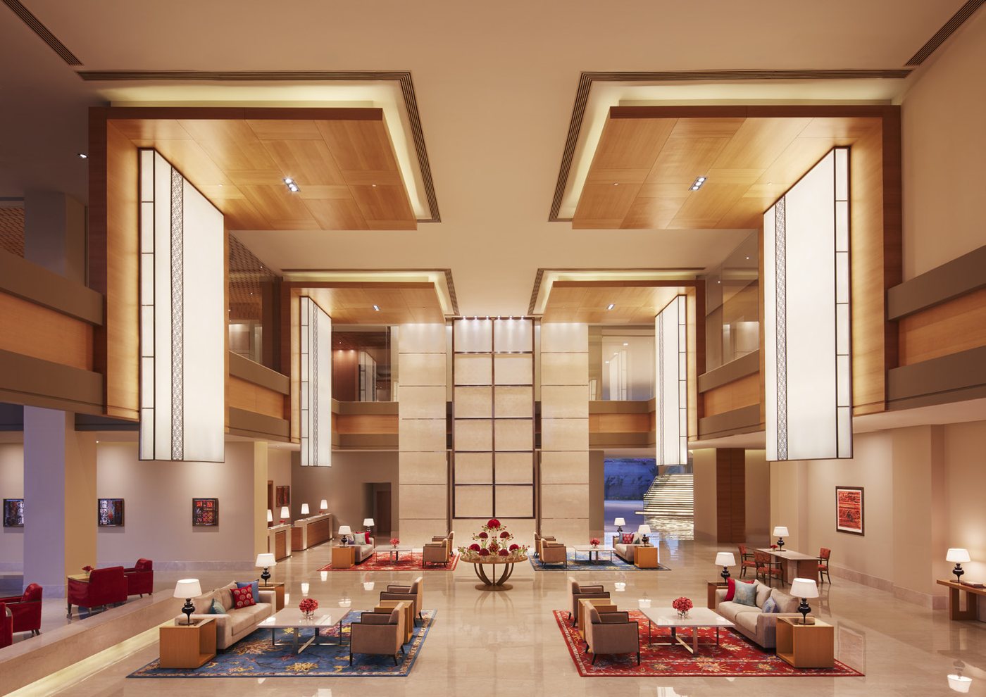 The double-height lobby at Taj Bangalore — pale stone walls and timber-panelled soffits with four large square light boxes hanging over a polished marble floor, low sofas and armchairs on red and blue rugs, and a lit staircase to a water feature at the far end