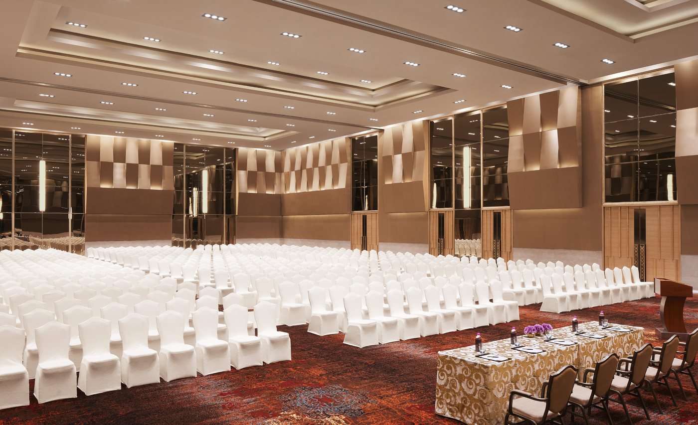 The Grand Ballroom at Taj Bangalore set theatre-style — several hundred white-covered chairs in rows facing a gold-skirted head table and lectern, under a deeply coffered white ceiling, with bronze mirrored and folded-metal wall panels down both sides and a patterned deep-red carpet
