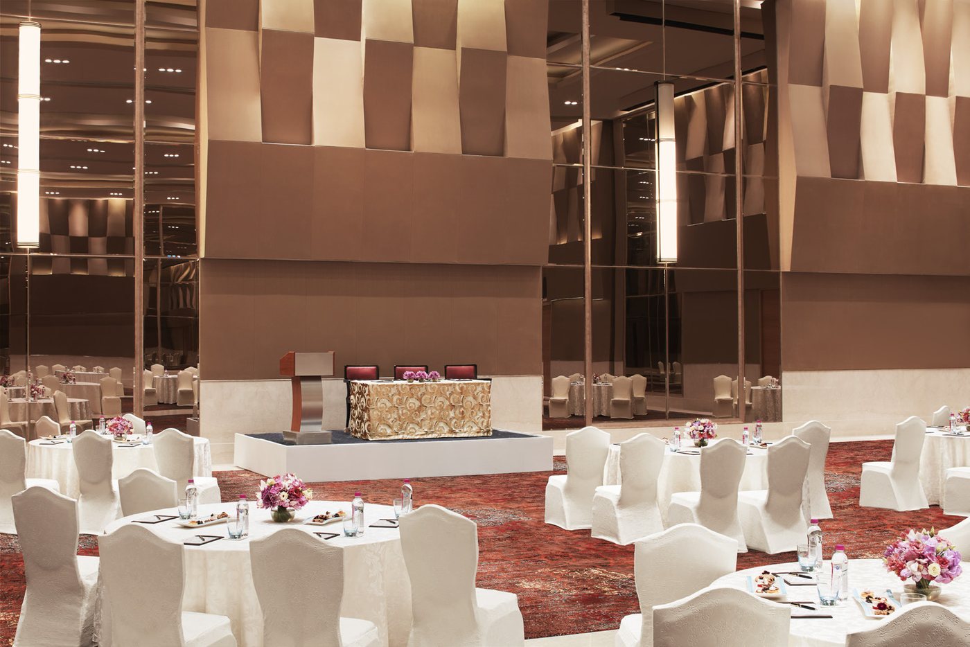 The Grand Ballroom at Taj Bangalore laid with round tables in white covers and pink floral centrepieces facing a low white stage with a gold-patterned head table, tall cylindrical light columns and bronze folded-metal wall panels behind