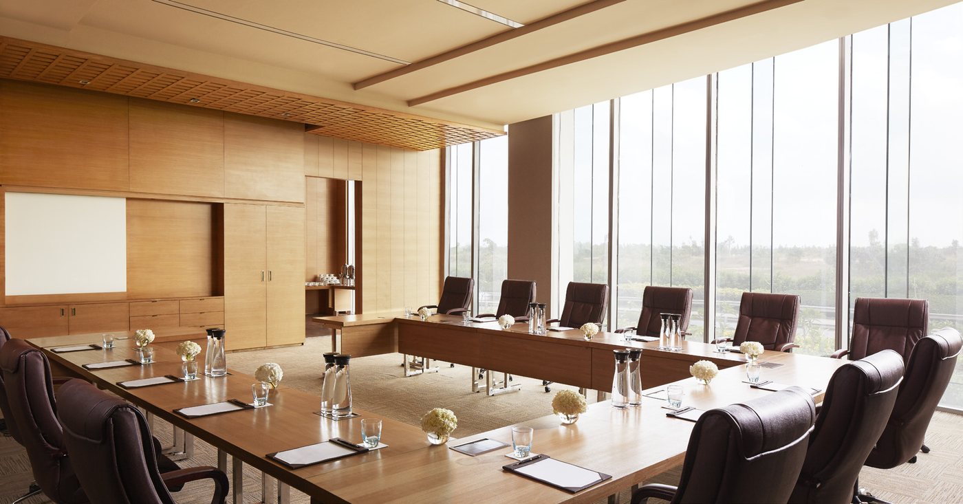 A daylit meeting room at Taj Bangalore set in a hollow square — timber-veneer walls and ceiling, leather executive chairs at pale wood tables with water carafes and white hydrangeas, and a full-height window wall on the right looking out over trees and the airport boulevard