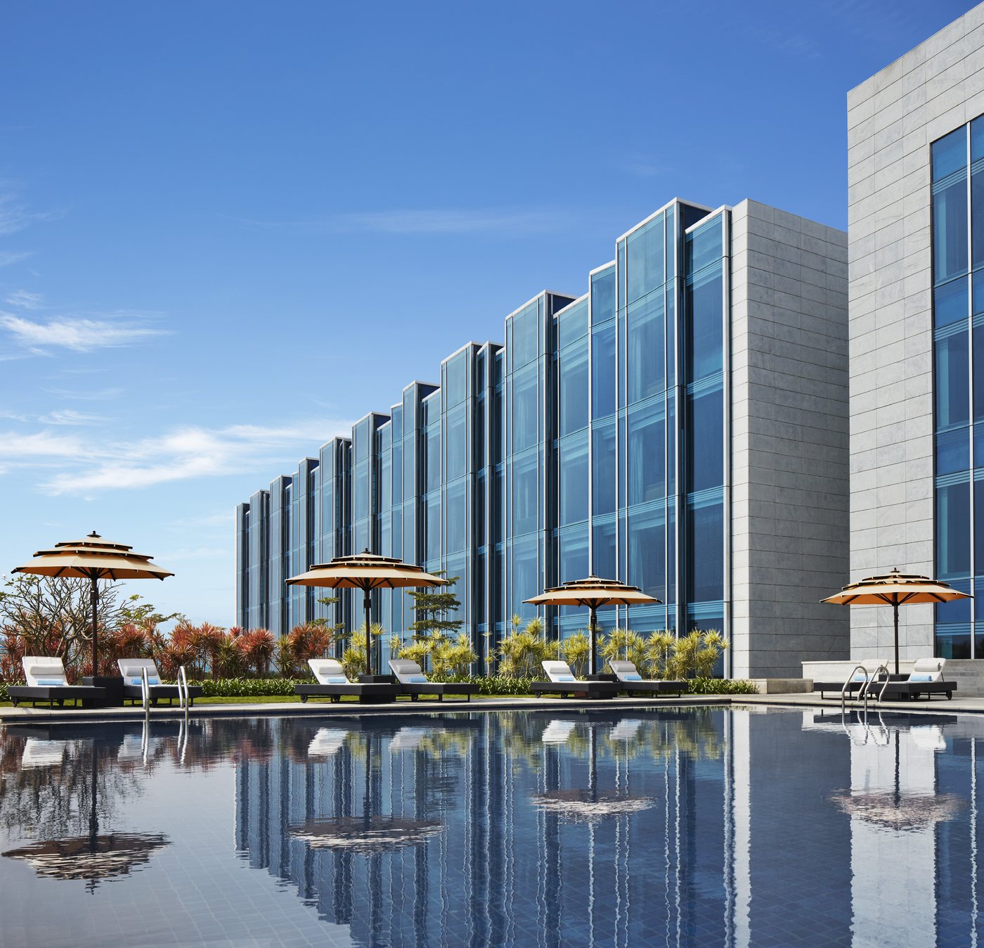 The outdoor pool at Taj Bangalore — a long rectangular pool reflecting the hotel's sawtooth blue-glass facade, with white loungers and tan parasols and a bed of red and yellow bromeliads along the far edge under a clear sky