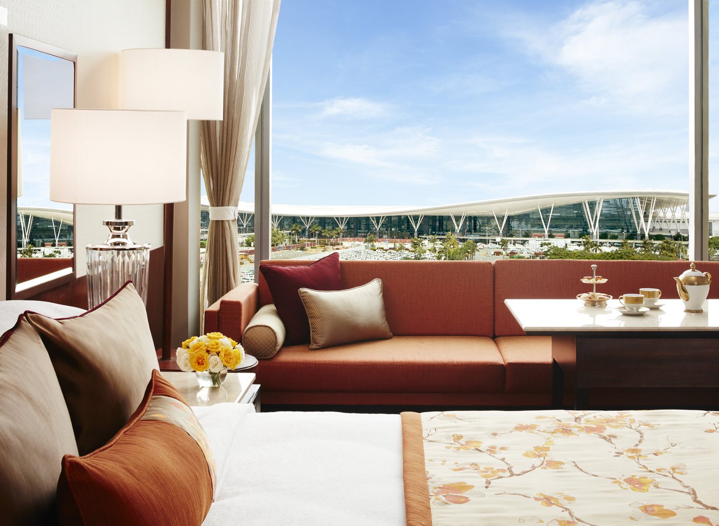 A guest room at Taj Bangalore looking out through a full-height window at the wave-roofed Kempegowda International Airport terminal across the car park — a burnt-orange banquette sofa and marble table by the glass, a crystal lamp and a bed with a cherry-blossom throw in the foreground