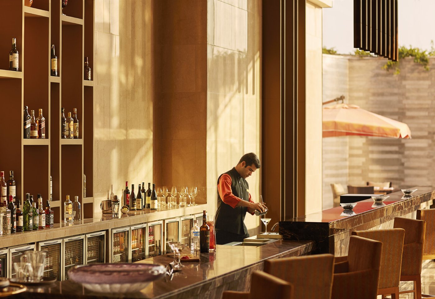 The bar at Taj Bangalore in late-afternoon light — a long marble and red-lacquer counter with a bartender mixing a drink, backed by open shelving of spirits, with an open-air terrace and an orange parasol visible through the glass beyond