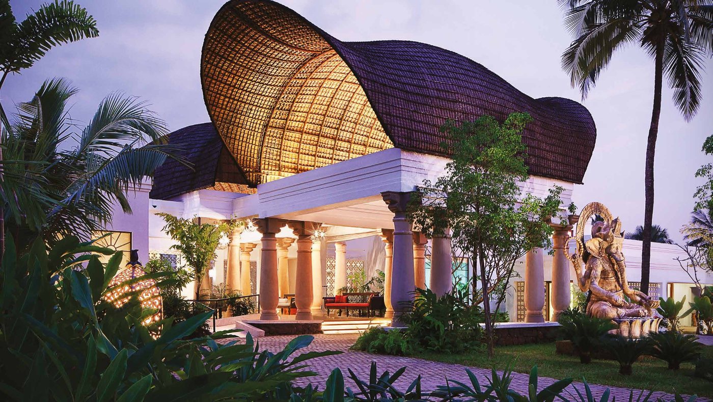 The arrival pavilion at Taj Bekal at dusk, a sculpted thatch canopy sweeping over the columned porte cochère with planting and palms around it
