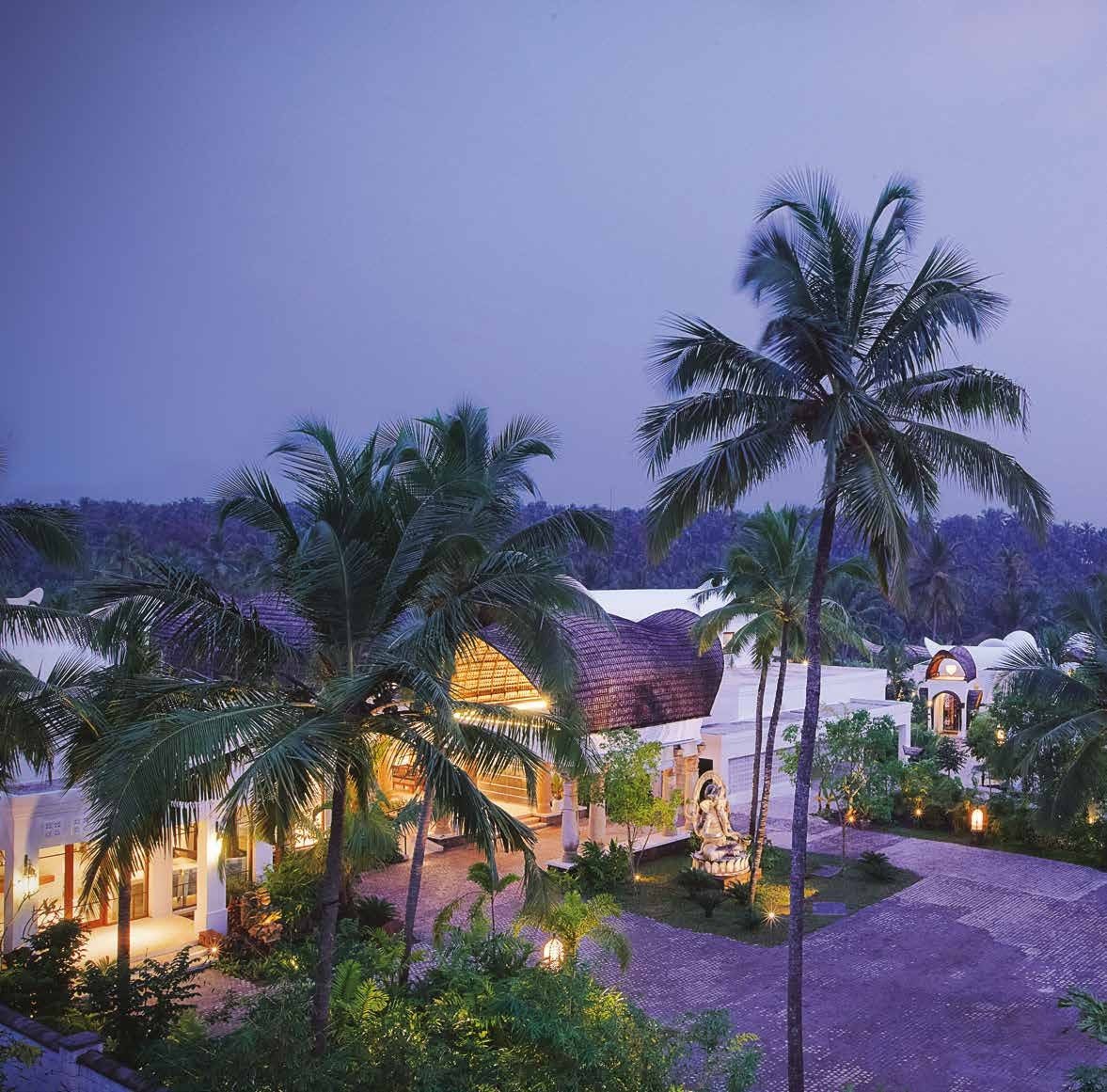 Taj Bekal at dusk seen through coconut palms, the low tiled roofs of the resort lit against a violet sky