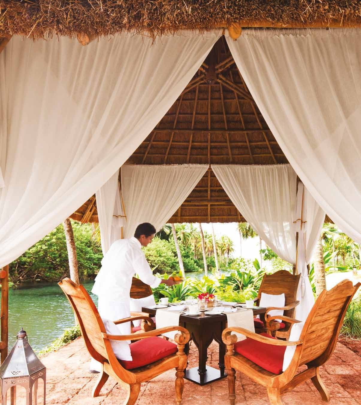 Thatched dining cabana beside the backwater at Taj Bekal, white drapes tied back and a waiter laying a table for two