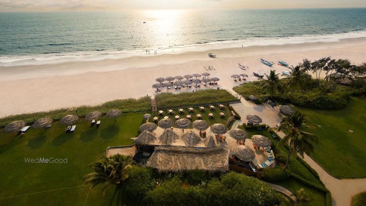 Aerial of Taj Exotica Goa's beach at sunset — the thatched beach restaurant and palm umbrellas at the lawn's edge, the sand and the Arabian Sea beyond