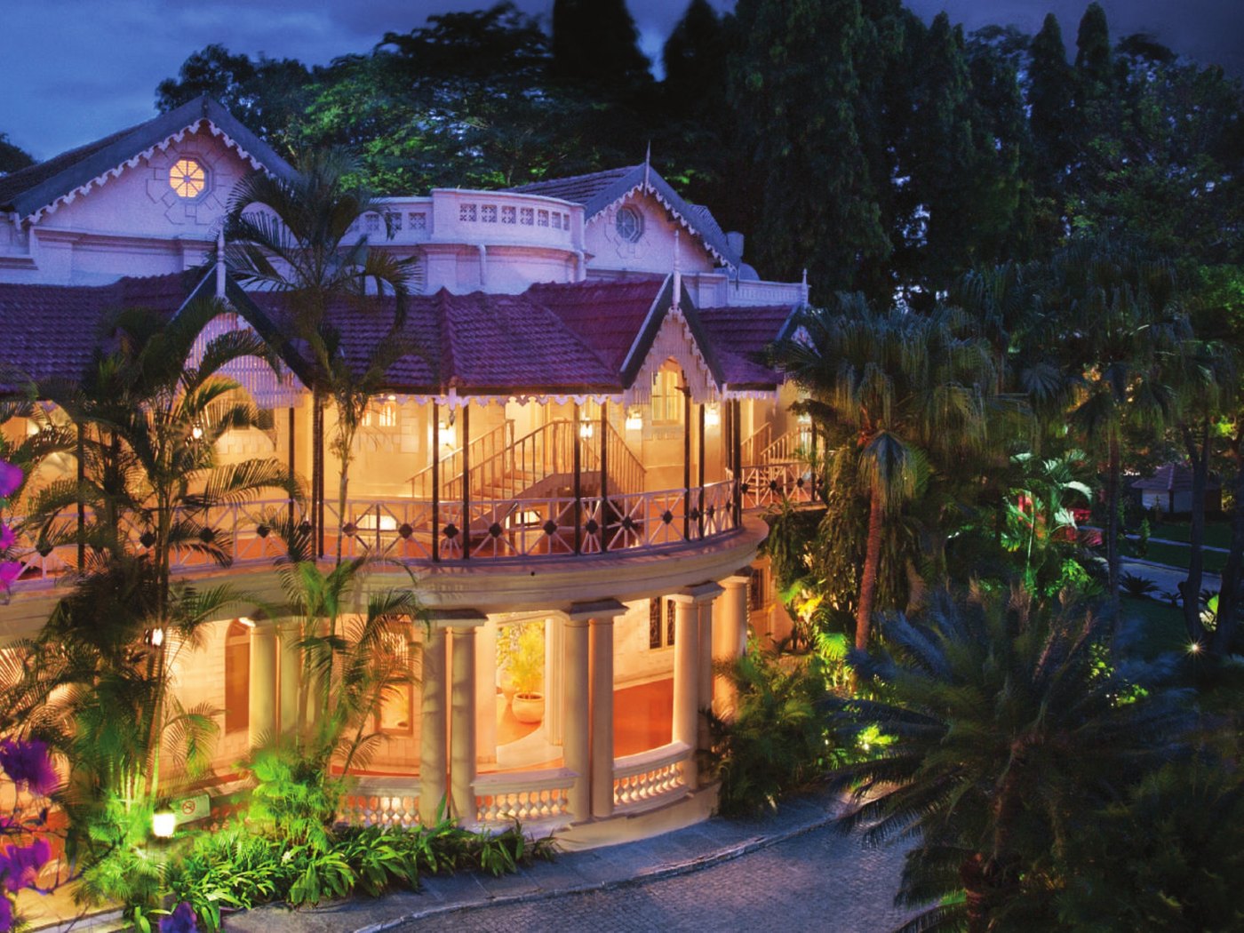 A heritage block at Taj West End Bengaluru floodlit at dusk — a two-storey colonial bungalow with a red-tiled roof, fretwork gables, a curved first-floor verandah on slim iron columns and a colonnaded ground floor, framed by palms and bougainvillea over the driveway