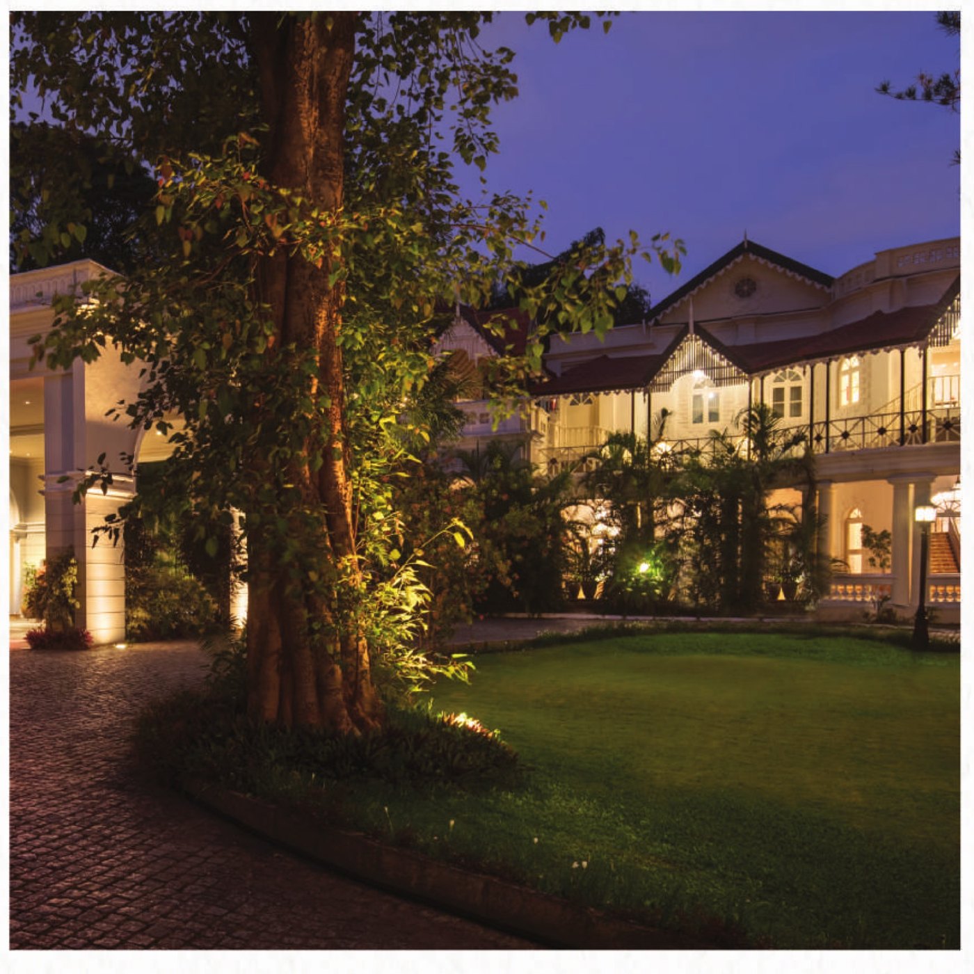 Evening view across a lit lawn at Taj West End Bengaluru — a large uplit tree in the foreground, a brick-paved drive curving past a white colonnaded porch, and the long two-storey heritage wing with tiled gables and iron verandah railings glowing behind