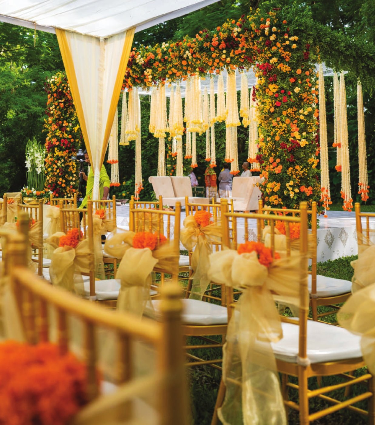 A wedding ceremony set on a lawn at Taj West End Bengaluru under a white marquee — a mandap banked in orange, yellow and red roses with long cream floral strings hanging from the canopy, two cream chairs on a low stage, and gold chiavari chairs tied with gold organza bows and marigold posies in the foreground, garden trees behind