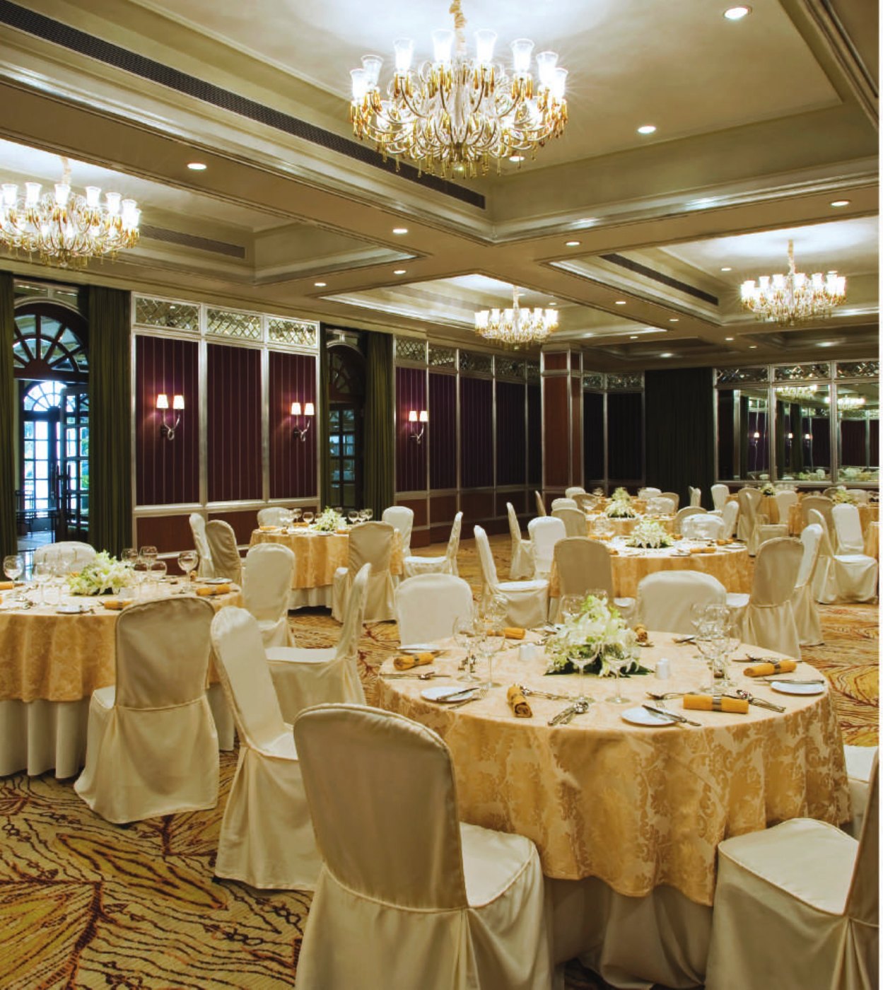 The ballroom at Taj West End Bengaluru set for a banquet — gold damask-topped round tables with cream chair covers and white floral centrepieces, three gold-and-white chandeliers on a coffered ceiling, mirrored and aubergine-panelled walls, an arched door to the garden at the left and a patterned gold carpet