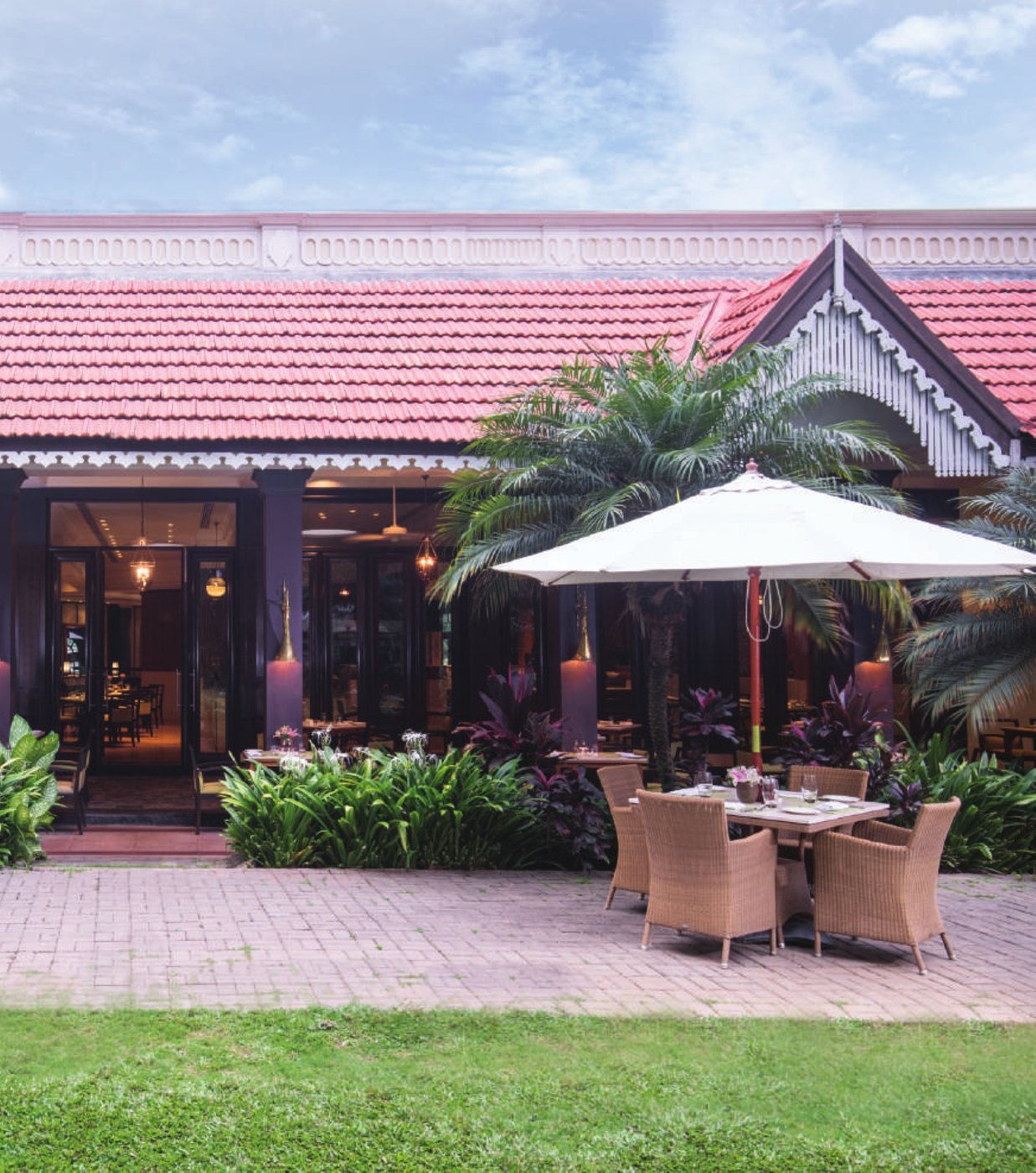 The open-air dining terrace beside Blue Ginger at Taj West End Bengaluru — a pink-tiled gabled roof with white fretwork over a black-framed pavilion, a cream parasol shading a set table with cane chairs on brick paving, tall palms and dense planting around it and lawn in the foreground