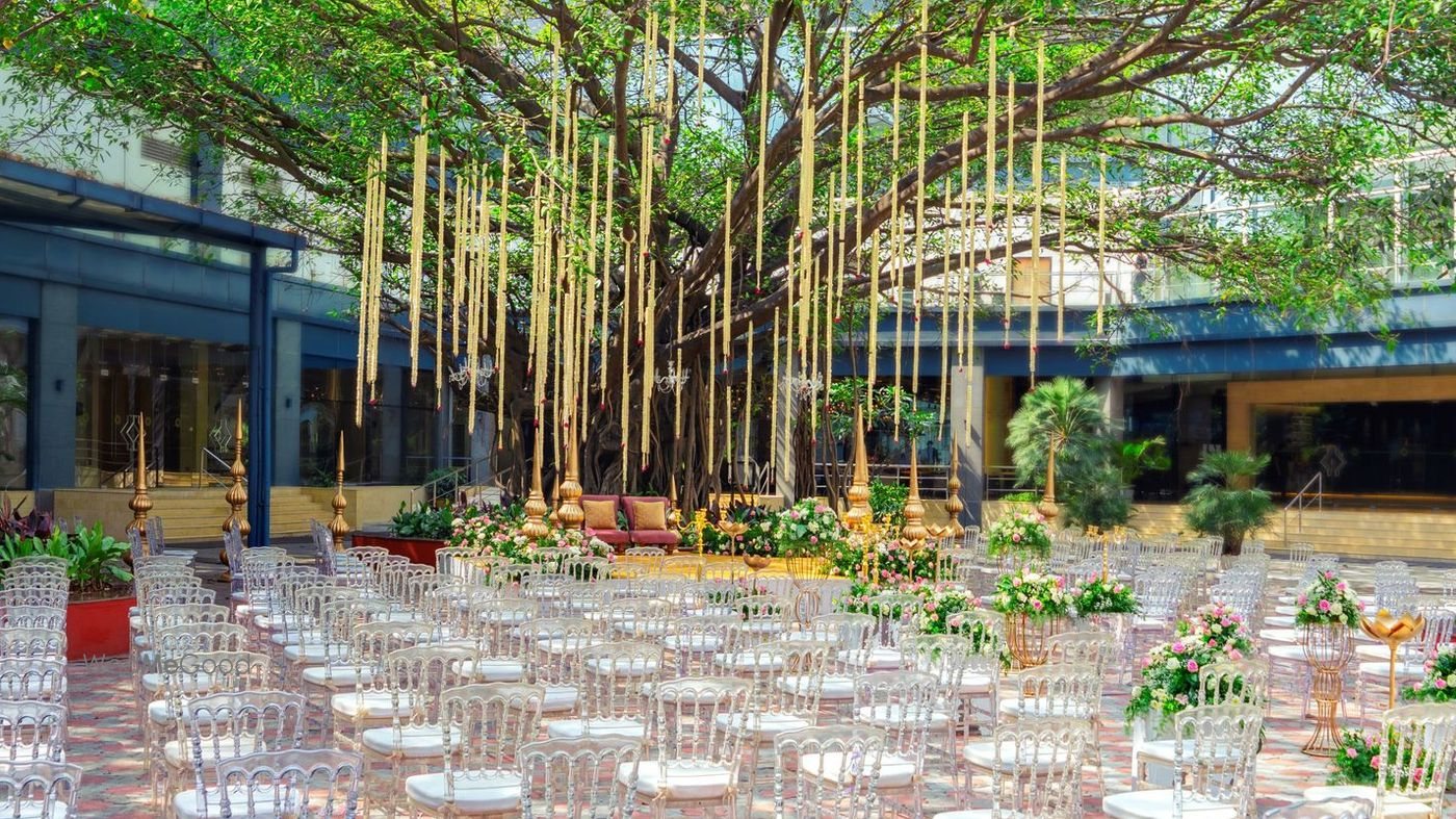 The banyan courtyard at Taj Yeshwantpur set for a ceremony — rows of clear chairs and low floral markers beneath a mature banyan tree, with slender poles rising into the canopy and the hotel facade behind