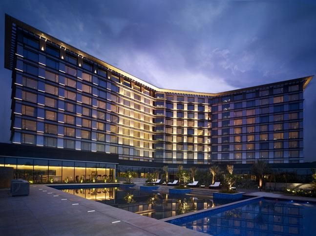 Taj Yeshwantpur exterior at dusk — the hotel's curved glass block lit from within above a long swimming pool and terrace