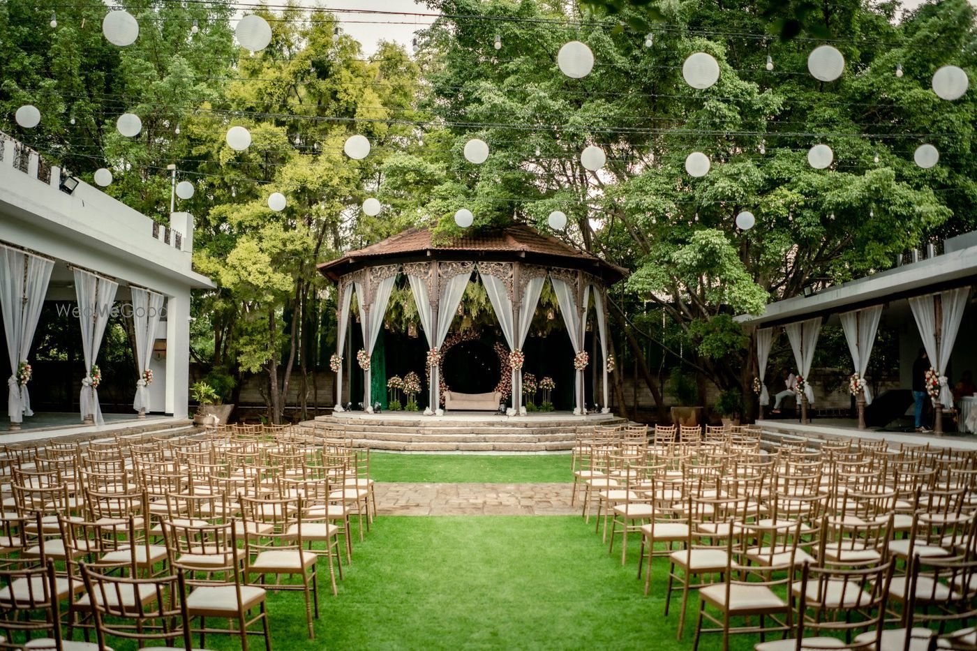 The colonial Bandstand at The Tamarind Tree — an open octagonal pavilion with fretwork columns, dressed in white drapes at the head of the lawn, with rows of gold chiavari chairs on both sides and white paper lanterns strung overhead
