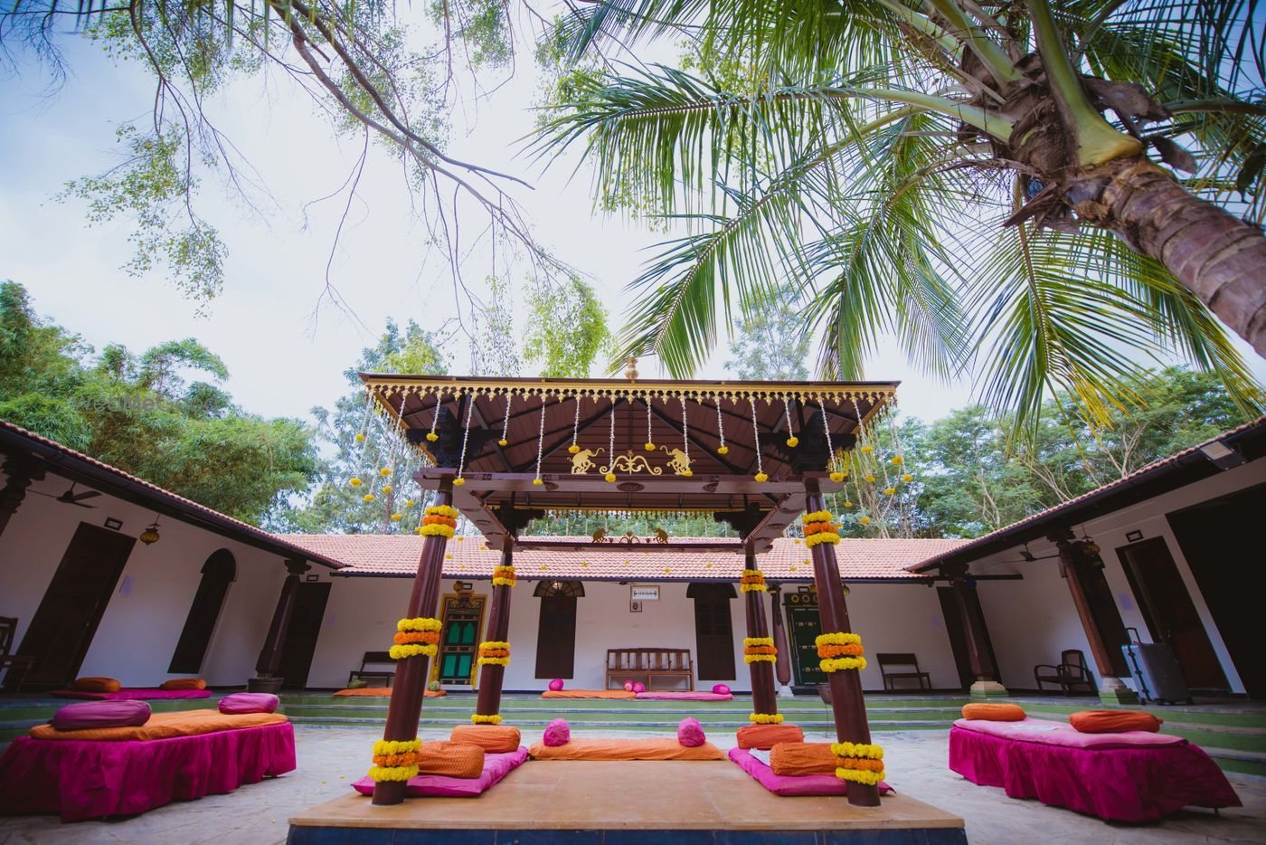 A carved wooden four-pillar mantapa at The Tamarind Tree hung with marigold and rose garlands, standing on a raised platform in a tiled-roof courtyard with pink and orange floor seating around it and coconut palms overhead