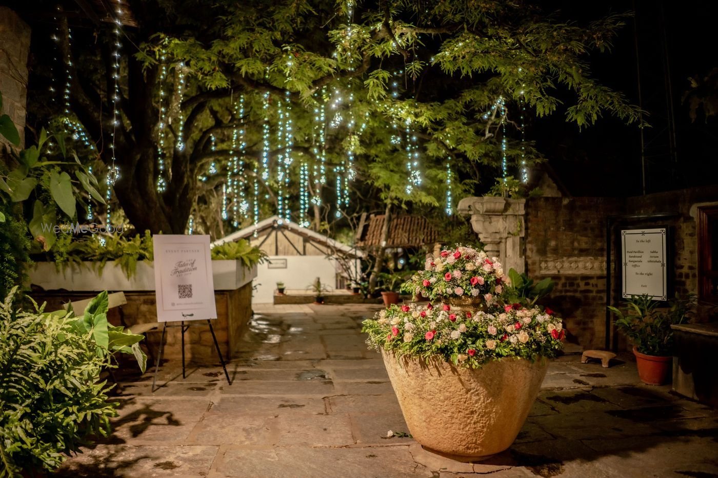 A stone-paved courtyard at The Tamarind Tree at night under a spreading tree strung with cascading fairy lights, a large terracotta urn of pink and white flowers in the foreground