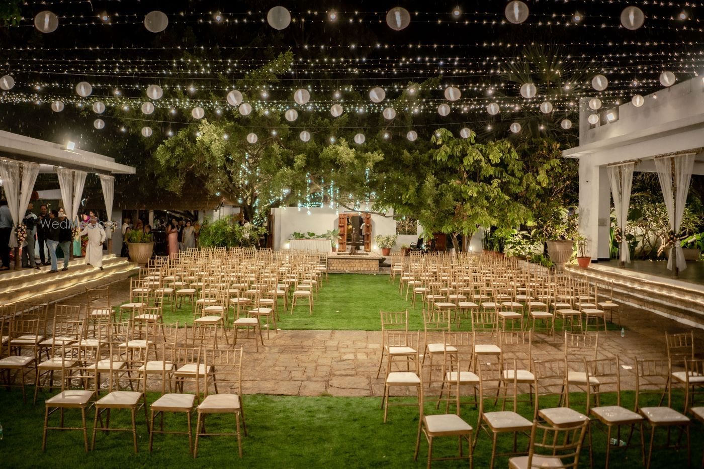 Rows of gold chiavari chairs set on lawn and stone paving at The Tamarind Tree facing a small white pavilion, under a canopy of paper lanterns and fairy lights strung between the estate buildings