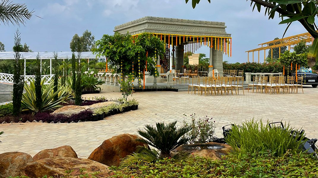 A stone-columned pavilion at Utsavaa above a rock garden — carved posts and a tiled roof with planted beds, boulders and a paved path running beneath it