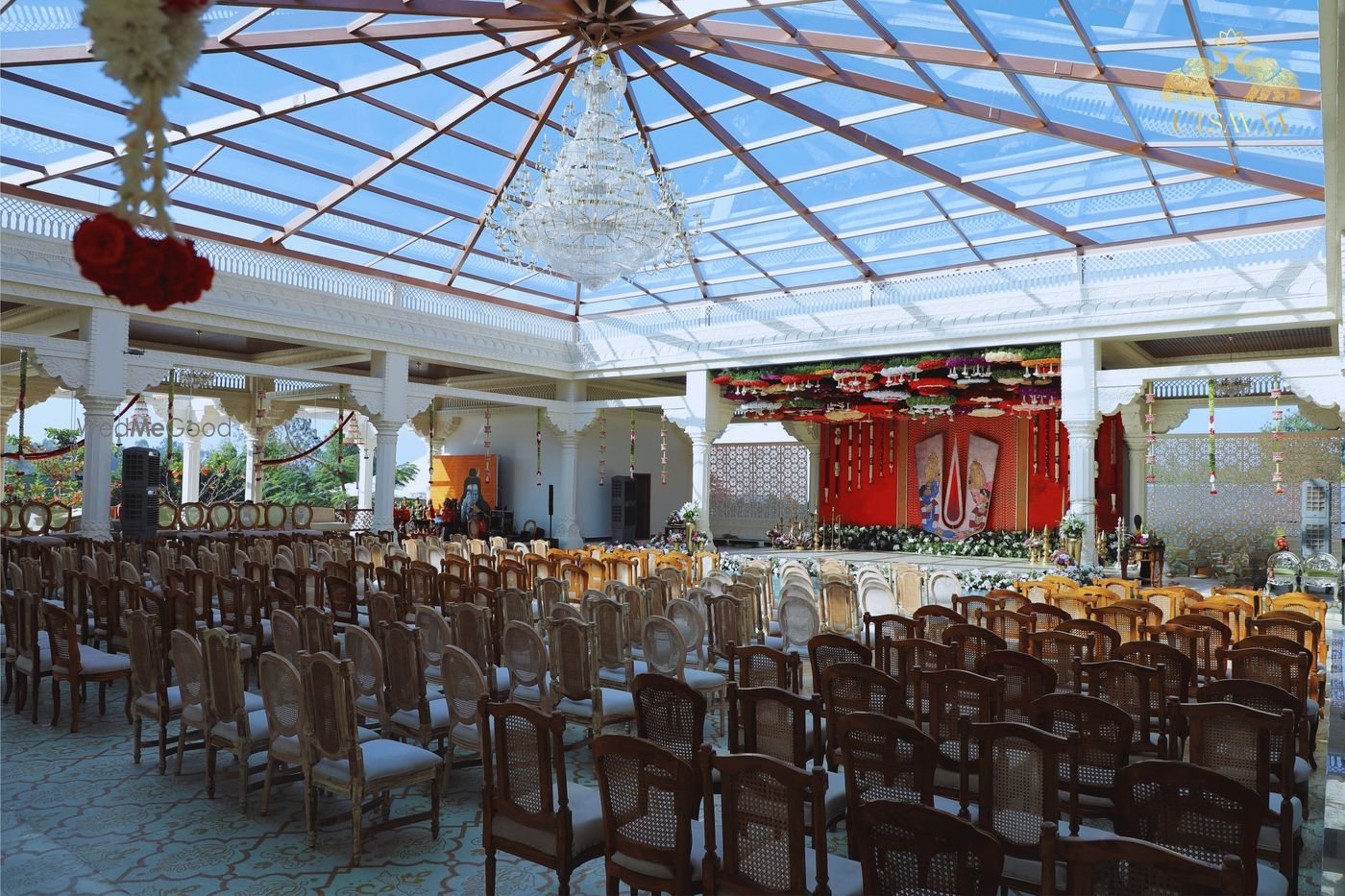 A ceremony pavilion at Utsavaa under a clear pitched roof — a red and gold draped mandap on a raised platform with a chandelier above and wooden chairs in rows