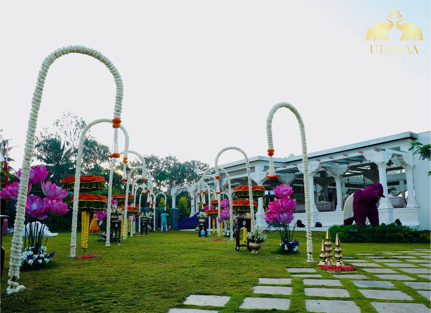 A lawn at Utsavaa dressed with white arch frames and pink floral trees on the grass, with a colonnaded white pavilion and a stone elephant at the far end