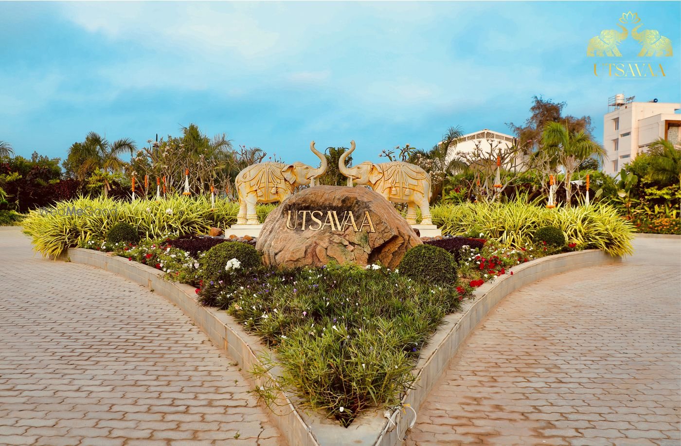 The entrance to Utsavaa — a carved stone name-block flanked by elephant sculptures in a landscaped roundabout, with the paved approach road running past