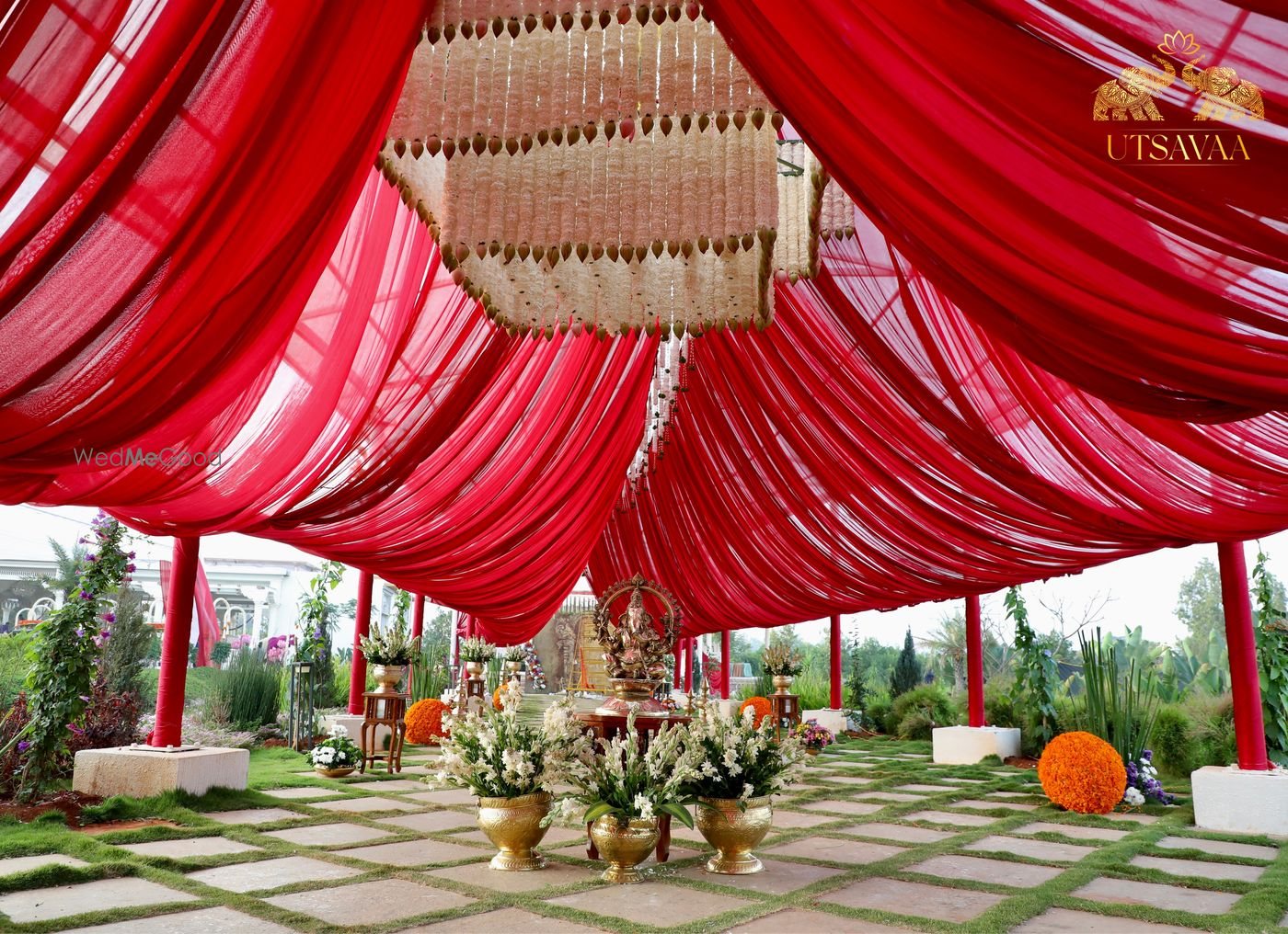 A ritual setting at Utsavaa under a deep red draped canopy on red columns, with brass vessels and marigold arranged on the stone platform beneath