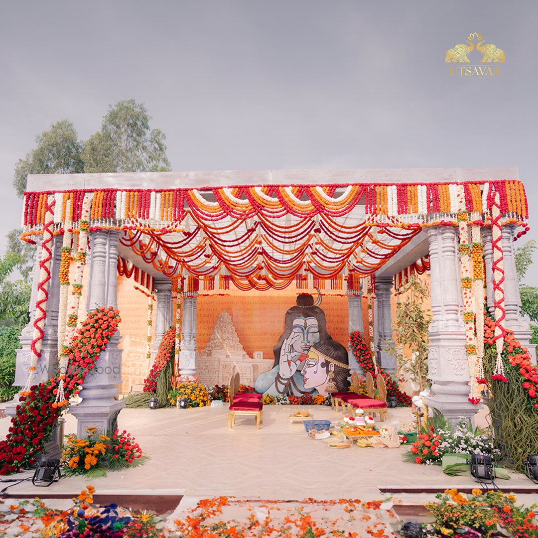 A carved stone mandapa at Utsavaa with a red and white striped canopy above it and a painted deity mural on the wall behind, floral garlands down the columns