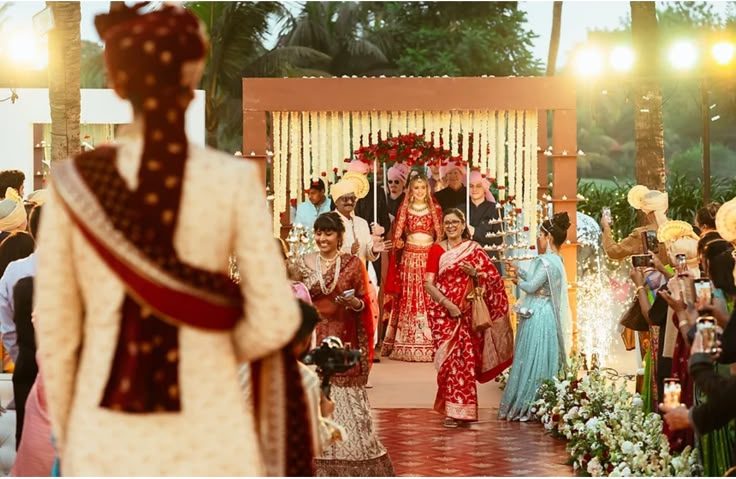 Bride's entry procession under a floral canopy towards the beach mandap, the groom waiting in the foreground