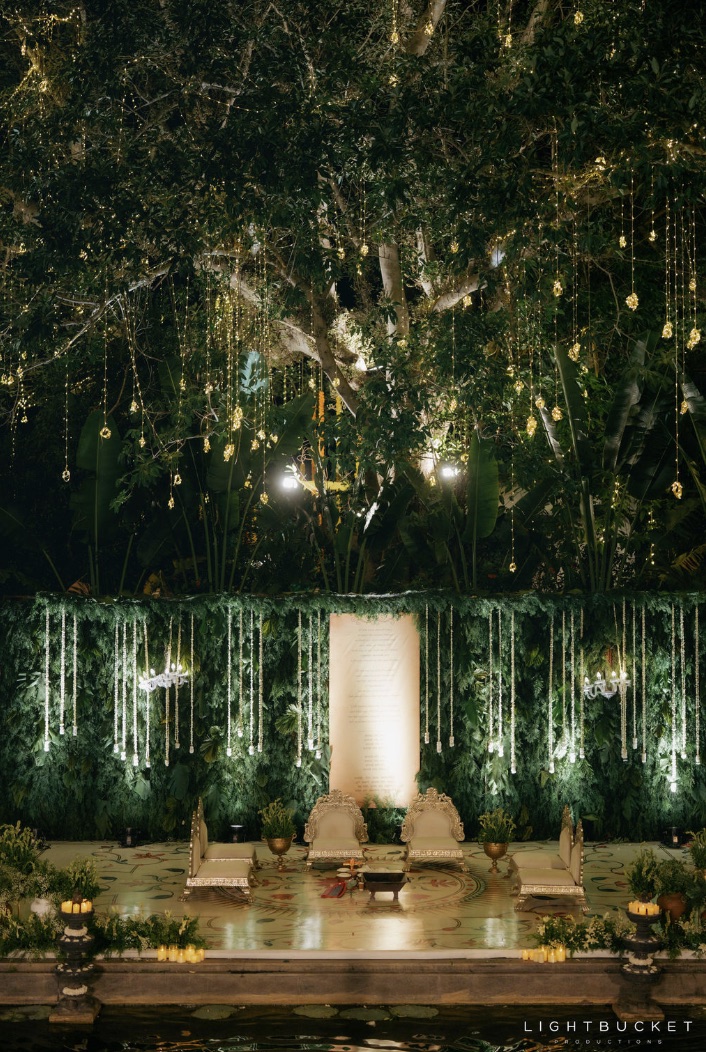 Greenery-wall wedding mandap with hanging white bloom strands, chandeliers and a printed vows scroll over the pond at Nature Knots Bangalore
