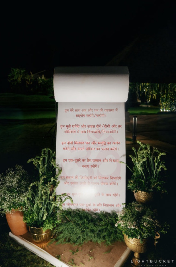 Freestanding scroll standee with wedding vows printed in Hindi among brass urns and plants on the lawn at Nature Knots Bangalore
