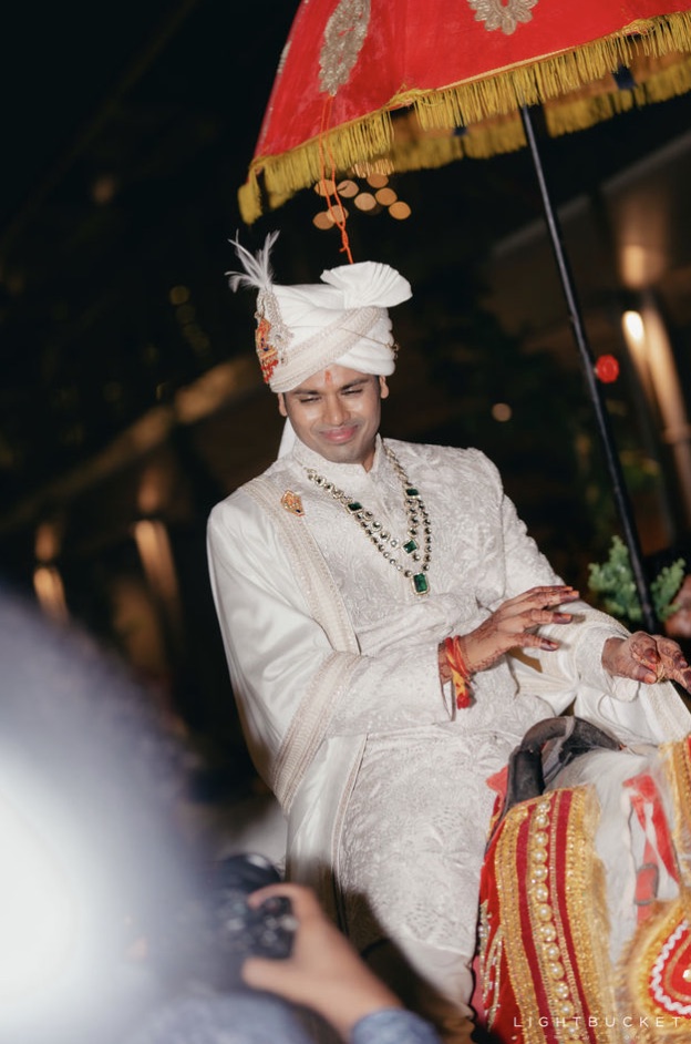 Groom in ivory sherwani on a mare under a red and gold embroidered baraat umbrella at Nature Knots Bangalore