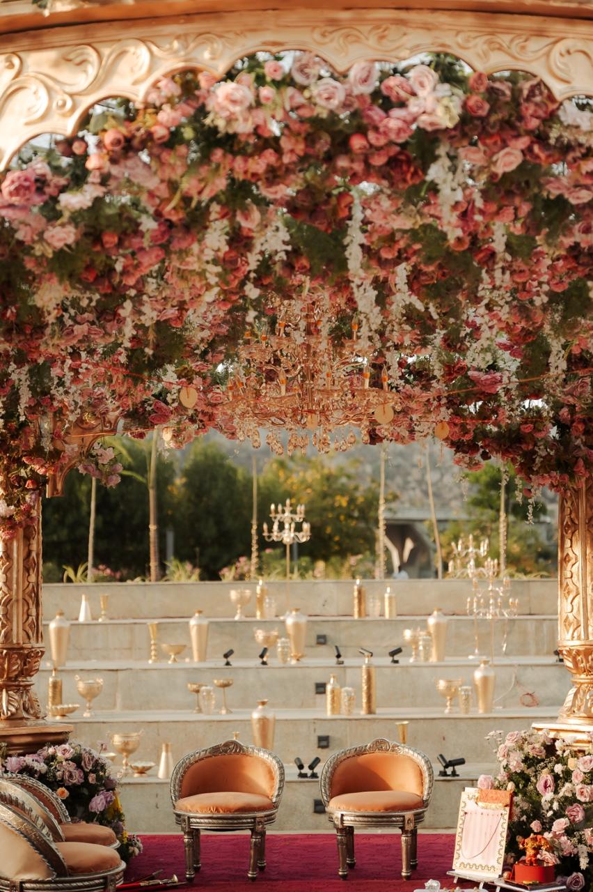 Floral ceiling of roses with a crystal chandelier inside the gold mandap at Amita Rasa, gold vessels on the amphitheatre steps behind