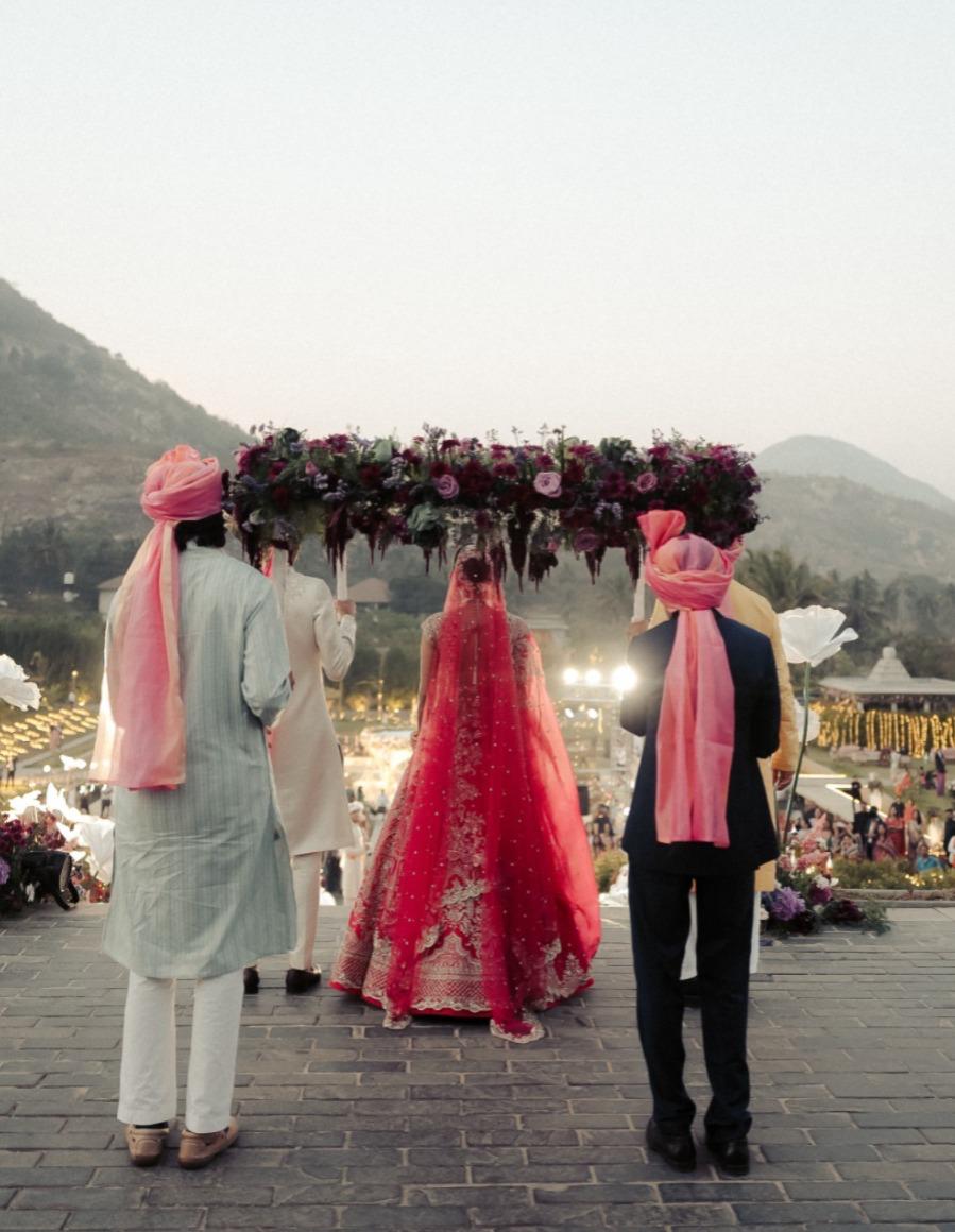 Bride in red lehenga walking under a burgundy floral chadar carried by men in pink safas at Amita Rasa, Nandi Hills near Bangalore