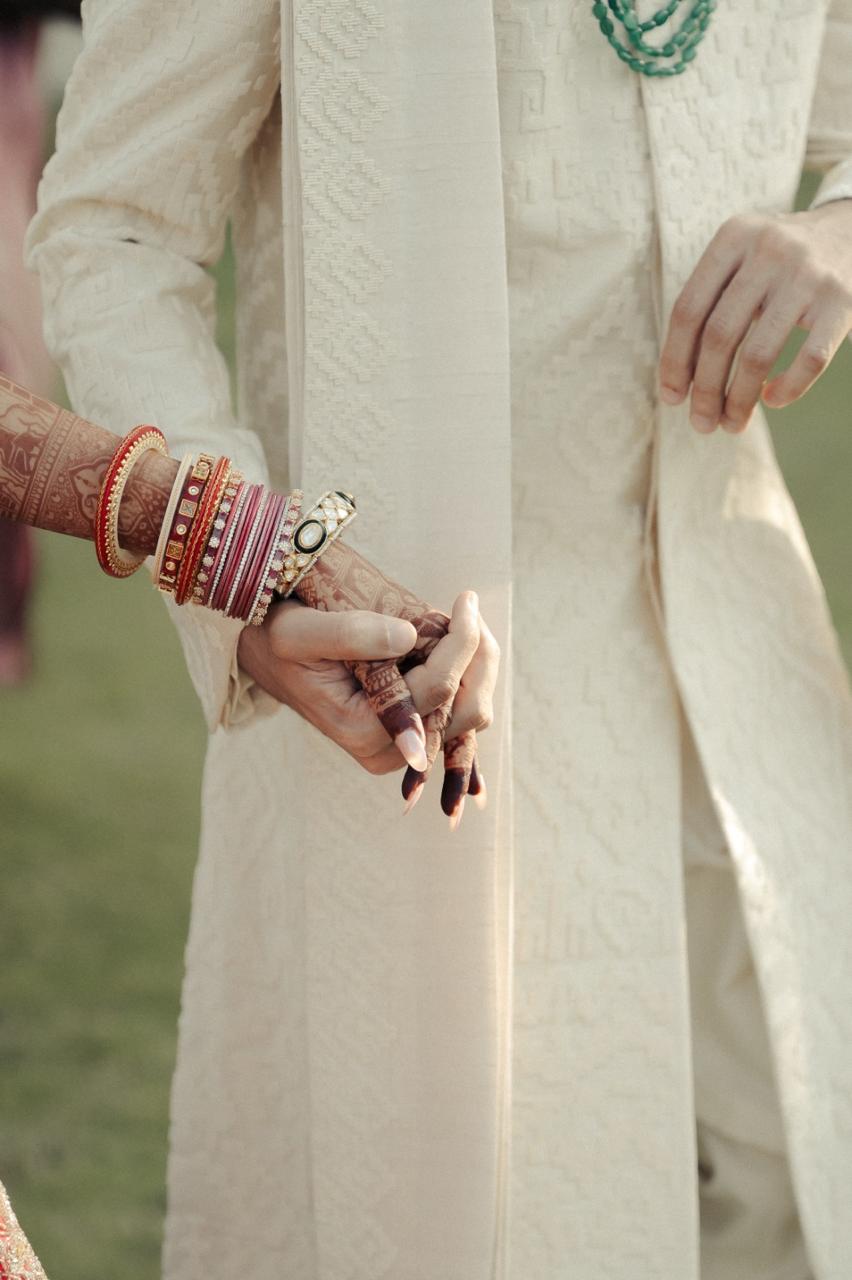 Bride's hands in red chooda and mehendi holding the groom's hand against his ivory sherwani