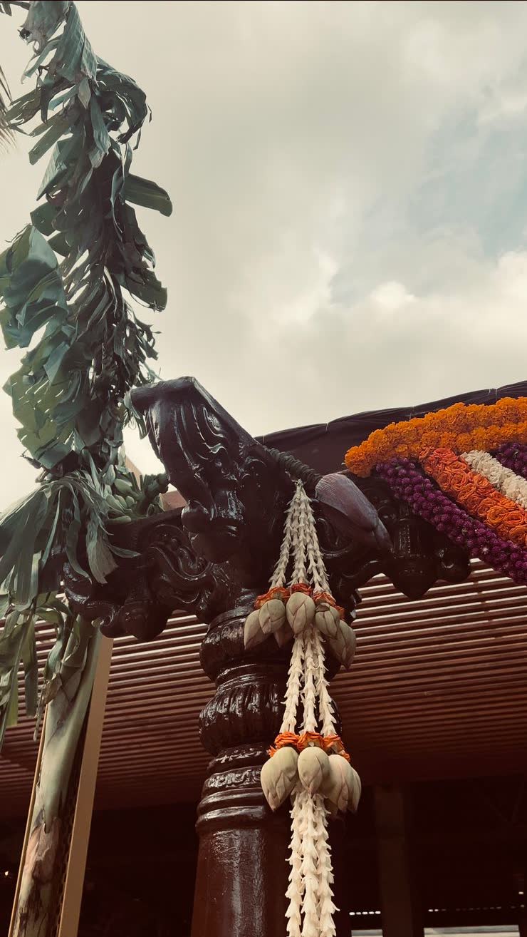 Carved dark wood column dressed with white lotus-bud garlands, shredded banana leaf toran and marigold and purple flower ropes against the sky